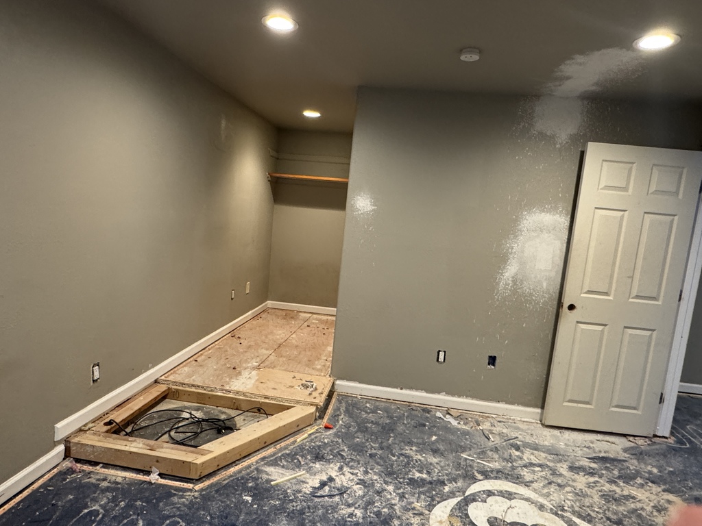 An empty room under renovation shows gray walls with visible patch marks, recessed ceiling lights, and a partially removed raised platform exposing wooden framing and cables. A white door stands open on the right, and construction dust and debris cover the floor, indicating ongoing remodeling work.