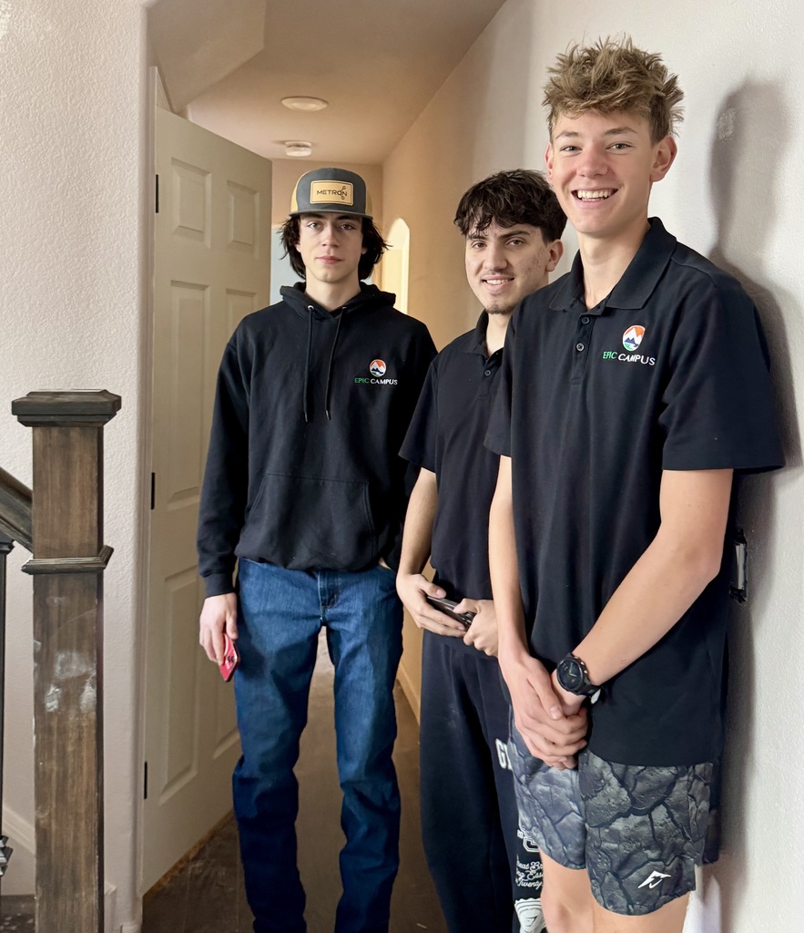 Three high school students stand in a hallway inside a home. Two students wear black EPIC Campus shirts, one smiling at the camera, while another student in a black hoodie and baseball cap stands beside them holding a red phone. A white door and neutral-colored walls create a simple indoor background.