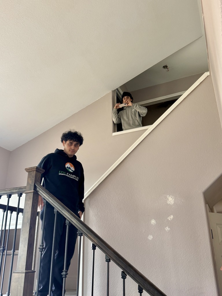 Two high school students stand inside a home stairwell. One student wearing a black EPIC Campus hoodie stands on the staircase landing, while another student in a gray sweatshirt and beanie leans out from an upstairs hallway opening, holding a phone as if taking a photo. Beige walls, a wooden stair railing with black metal balusters, and natural light from a nearby window frame the scene.