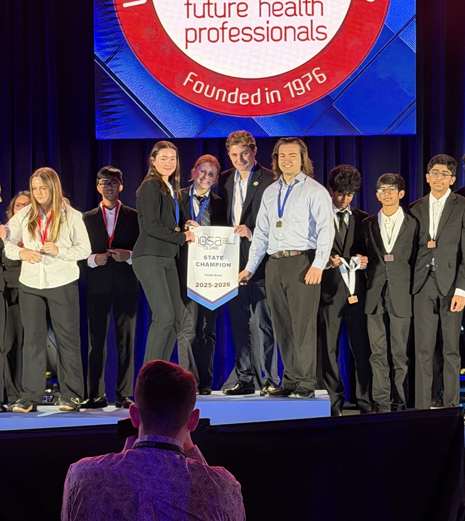 A group of high school students stand on a stage beneath a large screen reading “future health professionals – Founded in 1976.” Two students at center hold a banner that reads “HOSA State Champion 2025–2026.” Several students wear medals around their necks, dressed in formal business attire, as stage lighting highlights the award presentation.