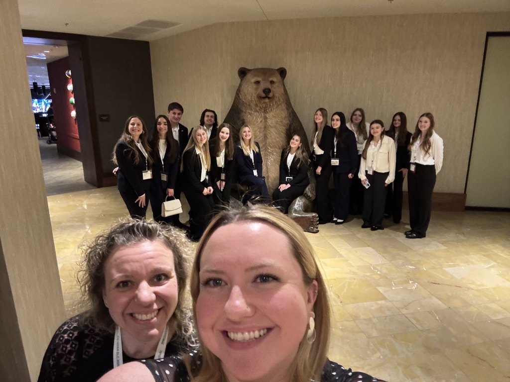 Two adult women smile in the foreground while taking a selfie in a hotel lobby. Behind them, a group of high school students in coordinated business attire pose around a large bear statue. The students wear conference badges, and the setting features beige walls and tiled flooring.