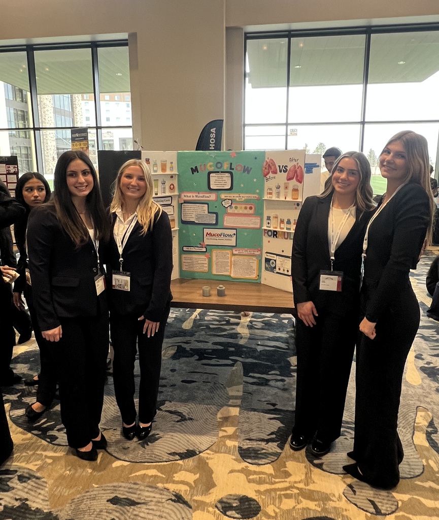 Four high school students dressed in black business attire stand on either side of a tri-fold presentation board titled “MucoFlow.” The board includes diagrams, text blocks, and labeled sections such as “Before vs After” with lung illustrations. The students wear conference name badges and stand in a large, well-lit venue with tall windows and patterned carpet.
