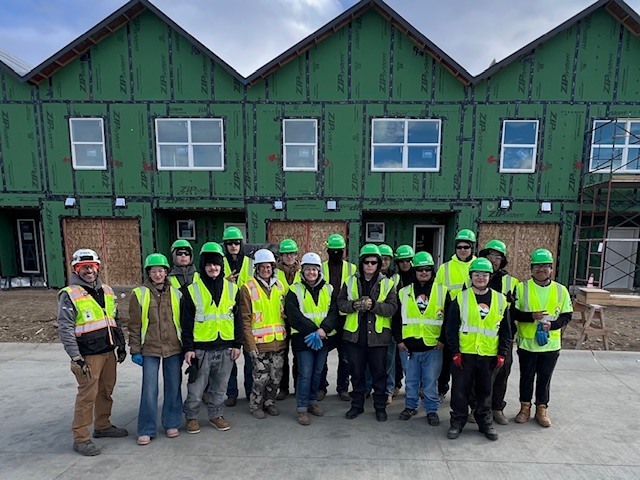 A group of students and mentors wearing bright safety vests and hard hats stand together for a photo in front of a partially constructed multi-unit building with green exterior sheathing and installed windows.