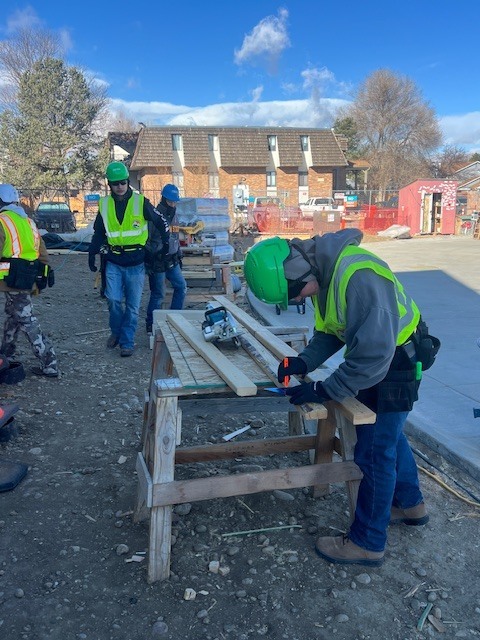 A student wearing a green hard hat, safety vest, gloves, and tool belt measures a board on a makeshift wooden workbench outdoors. Other students in safety gear walk behind them at the active construction site.