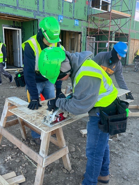 Two students wearing green hard hats and safety vests work together cutting a sheet of plywood on sawhorses with a circular saw, while another student stands nearby observing. A partially built house covered in green sheathing and scaffolding stands behind them.