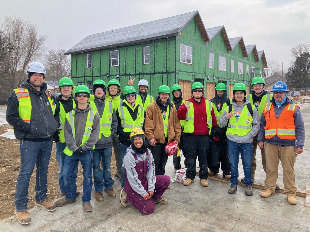 A diverse group of about fifteen students and adult mentors wearing green safety vests and hard hats pose together at a Habitat-style construction site in light snowfall. Behind them is a partially built multi-unit home covered in green sheathing with installed windows.