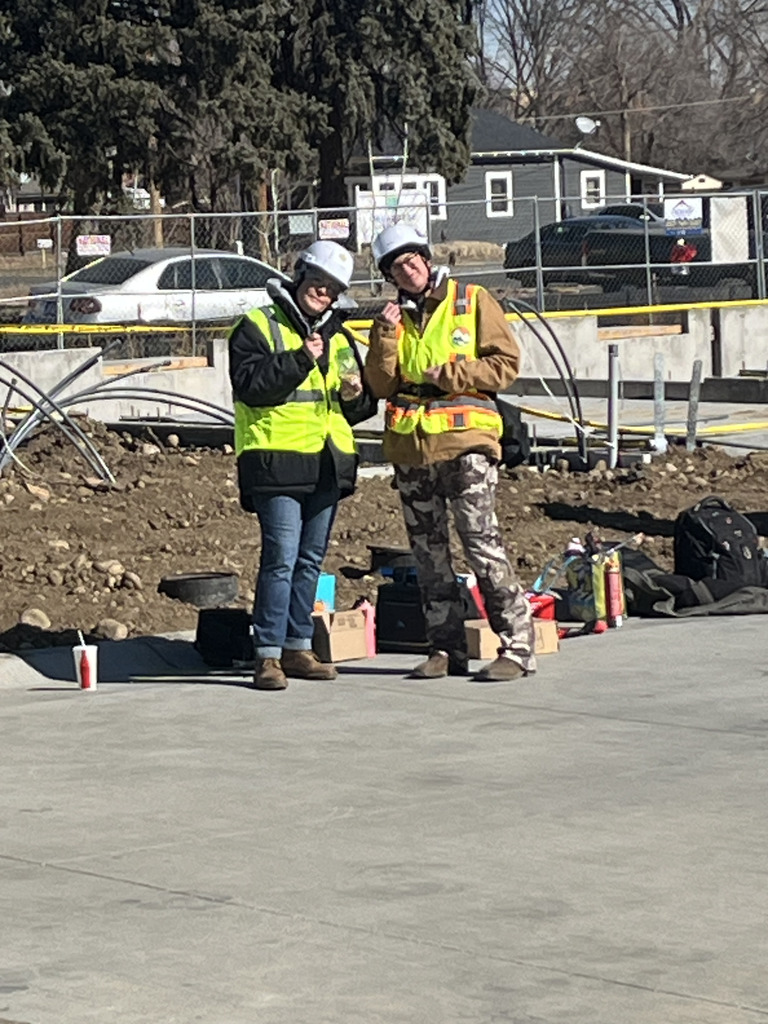Two students wearing white hard hats and bright safety vests smile and pose together on a concrete slab at a construction site. Tools, bags, and supplies are scattered behind them near a fenced area and nearby houses.