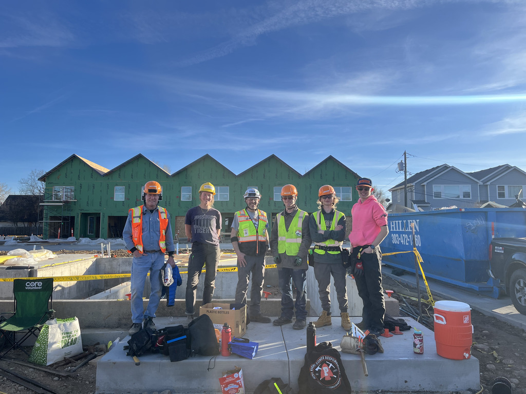 Six students and an adult wearing hard hats and safety vests stand together on a concrete foundation at a construction site. Tools, water bottles, and bags sit nearby, and a row of partially built homes with green exterior sheathing is visible in the background.