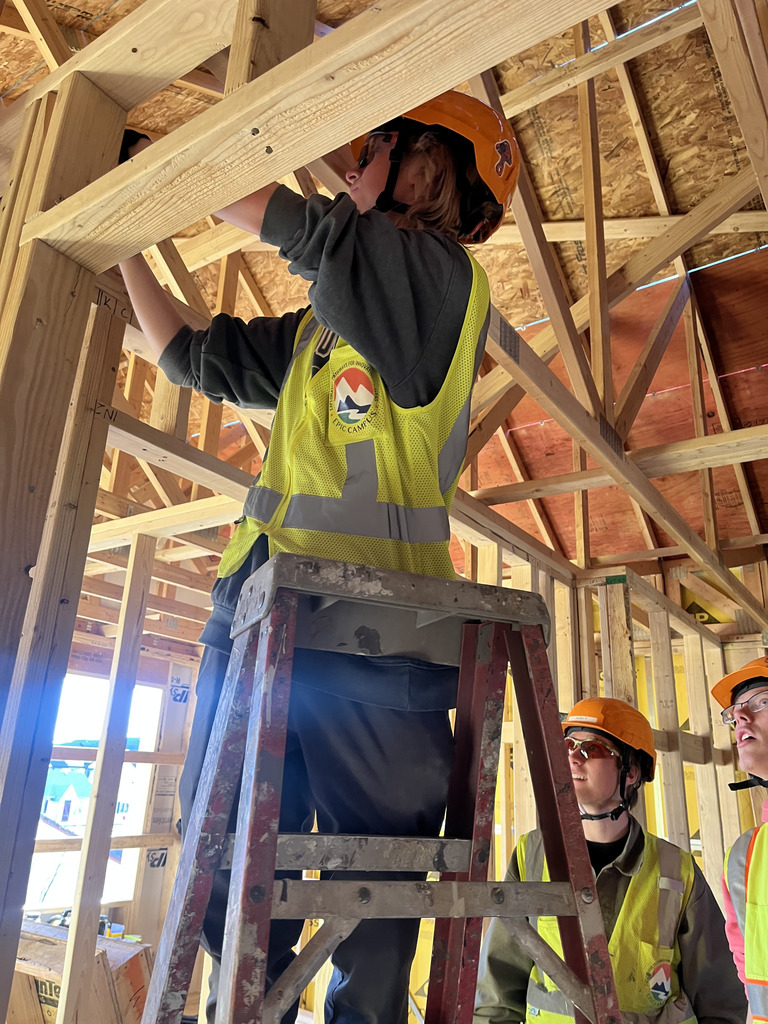 A student in an orange hard hat and bright safety vest stands on a step ladder inside a house under construction, reaching up to work on overhead framing. Two other students in safety gear watch from below inside the wood-framed structure.