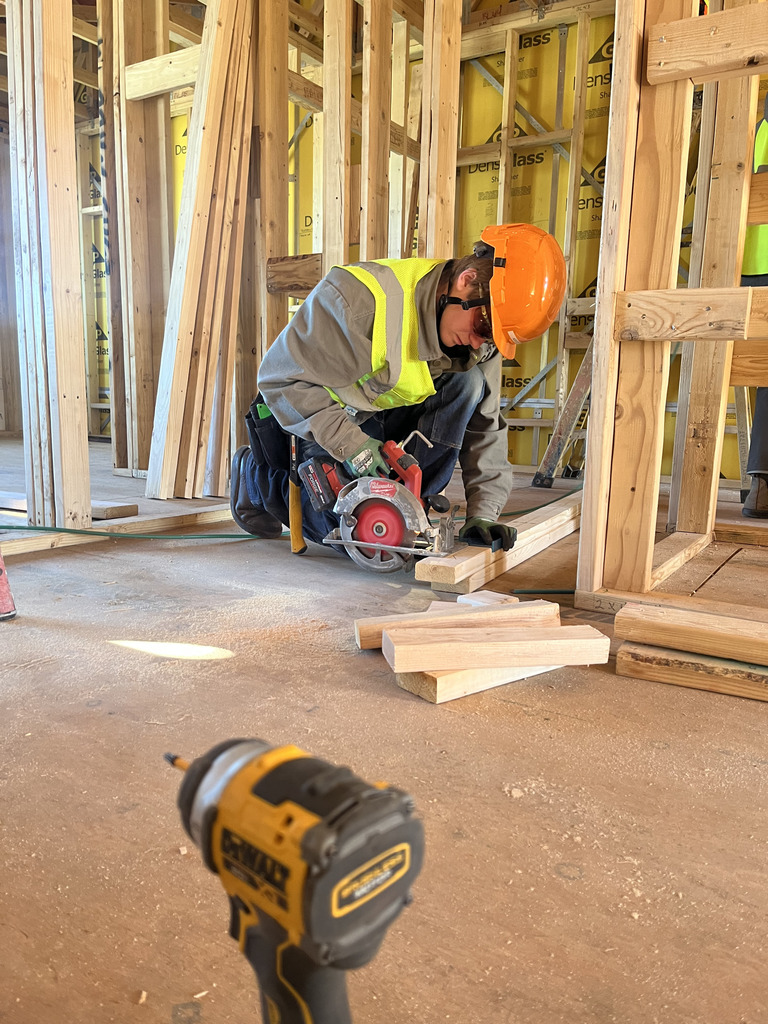 A student wearing an orange hard hat, safety glasses, and a bright safety vest kneels on a wooden floor inside a framed house structure, using a circular saw to cut a piece of lumber. Framing studs and yellow insulation sheathing are visible behind them, and a power drill rests in the foreground.