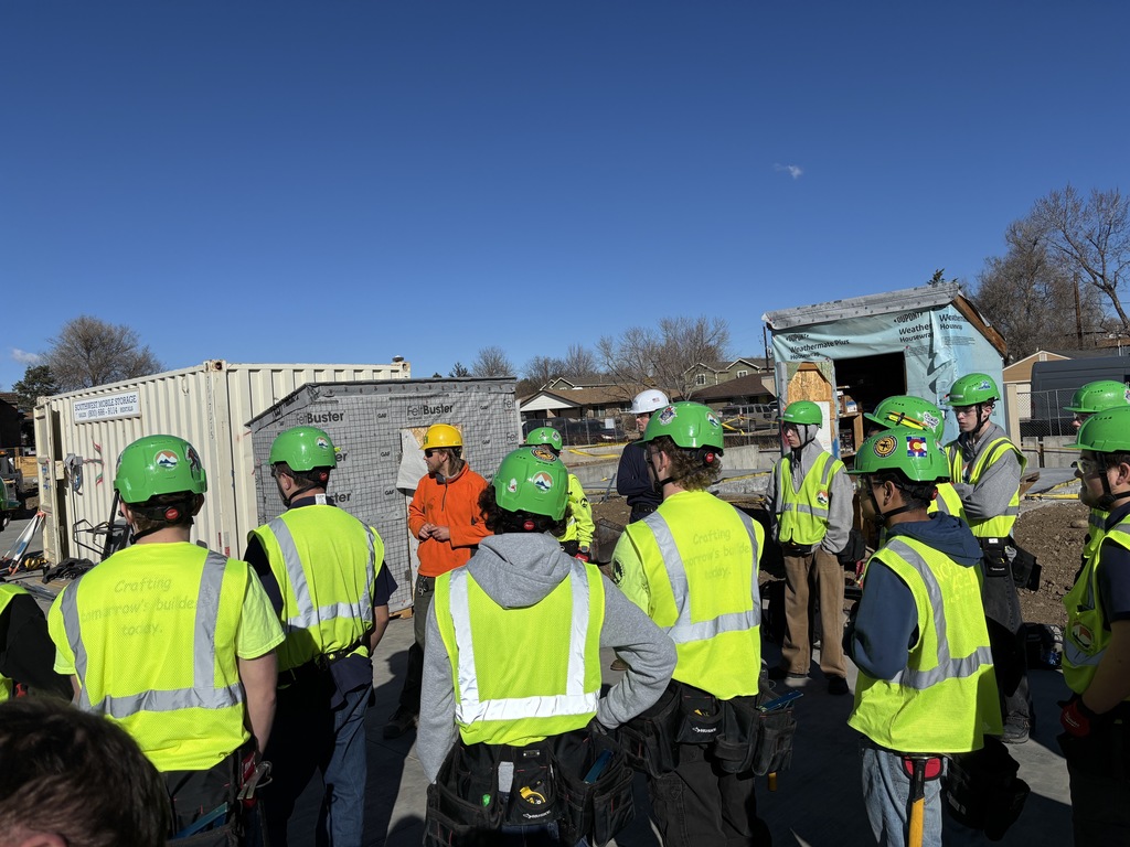 A group of students wearing green hard hats and bright safety vests stand in a semicircle listening to an instructor in an orange sweatshirt and yellow hard hat during a construction site briefing. Storage containers, equipment, and framed structures are visible behind them.