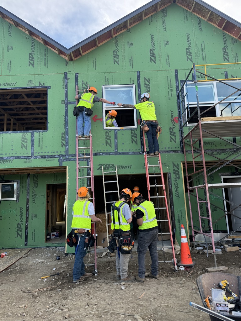 Several students wearing orange hard hats and bright safety vests work on ladders outside a partially built house. One student on a ladder passes a tool through a window opening to another student inside, while others stand below with tool belts and safety gear.