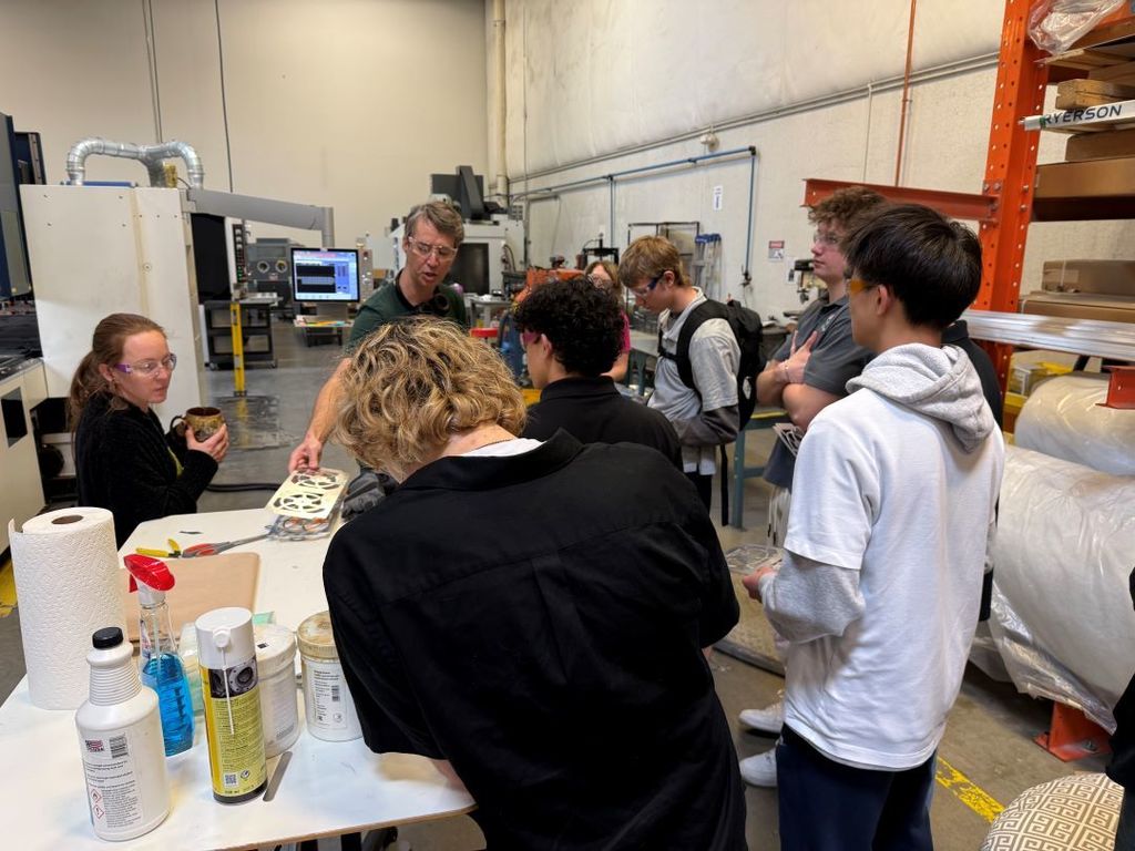 A small group of high school students gathers around two adults at a worktable in a manufacturing lab. One instructor holds a sheet of metal parts while explaining the process. The students—wearing casual clothing, backpacks, and safety glasses—stand closely watching the demonstration. Industrial machines, cleaning supplies, and storage shelves are visible around the workspace.