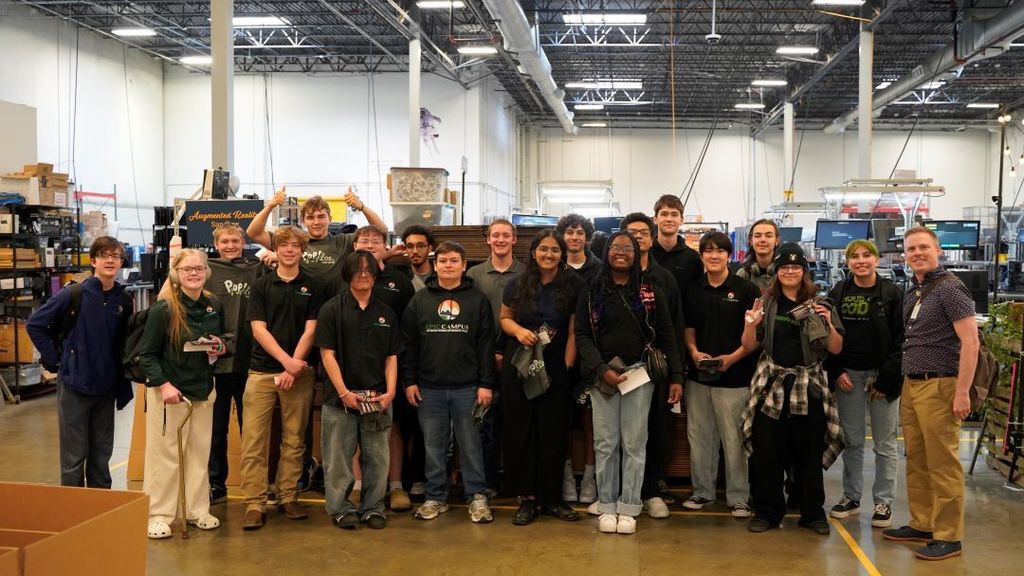 A large group of high school students and an adult chaperone stand together inside a spacious manufacturing or technology lab with high ceilings, exposed ductwork, and industrial equipment in the background. The diverse group of students—wearing a mix of school hoodies, polo shirts, and casual clothing—stand shoulder to shoulder smiling at the camera. Some students hold small items or bags from the visit. Workstations, machines, and shelves of materials fill the workshop behind them.