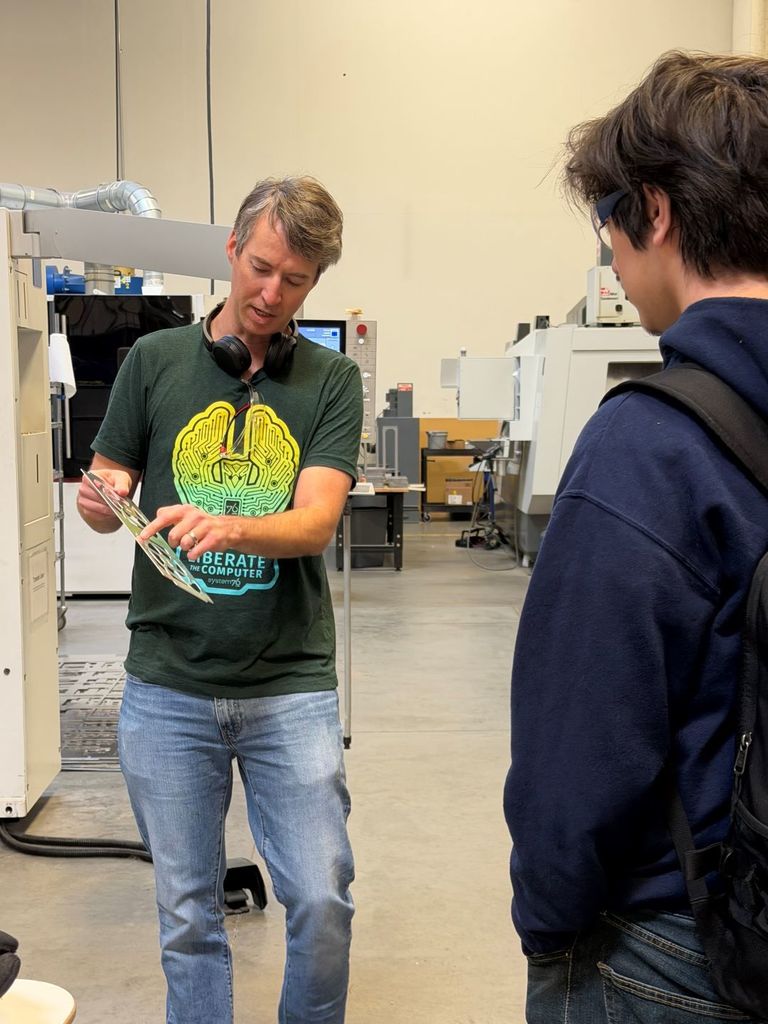 An adult instructor wearing a green T-shirt with a brain-and-circuit graphic and the words “Liberate the Computer – System76” holds and points to a thin metal panel with cut-out shapes while explaining it to a student. The student, wearing glasses, a navy hoodie, and a backpack, watches closely. They stand in a clean manufacturing space with large industrial machines and equipment visible in the background.