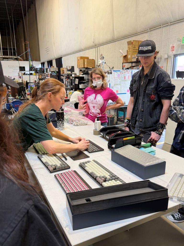 At a worktable inside a busy workshop, an adult staff member demonstrates assembling a mechanical keyboard while several high school students watch. Multiple keyboard cases, keycaps, and tools are spread across the table. A student in a pink T-shirt wearing a face mask holds a phone, while another student in a black jacket and cap observes nearby. Shelving, boxes, and workshop equipment fill the background.