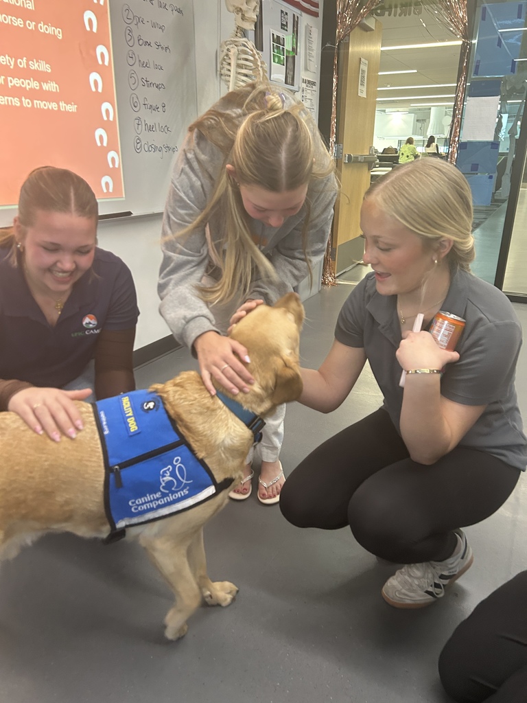 Three students crouch and smile while petting a yellow Labrador facility dog wearing a blue “Canine Companions” facility dog vest. The interaction takes place in a classroom with a projected slide, a whiteboard with notes, and a model skeleton in the background.