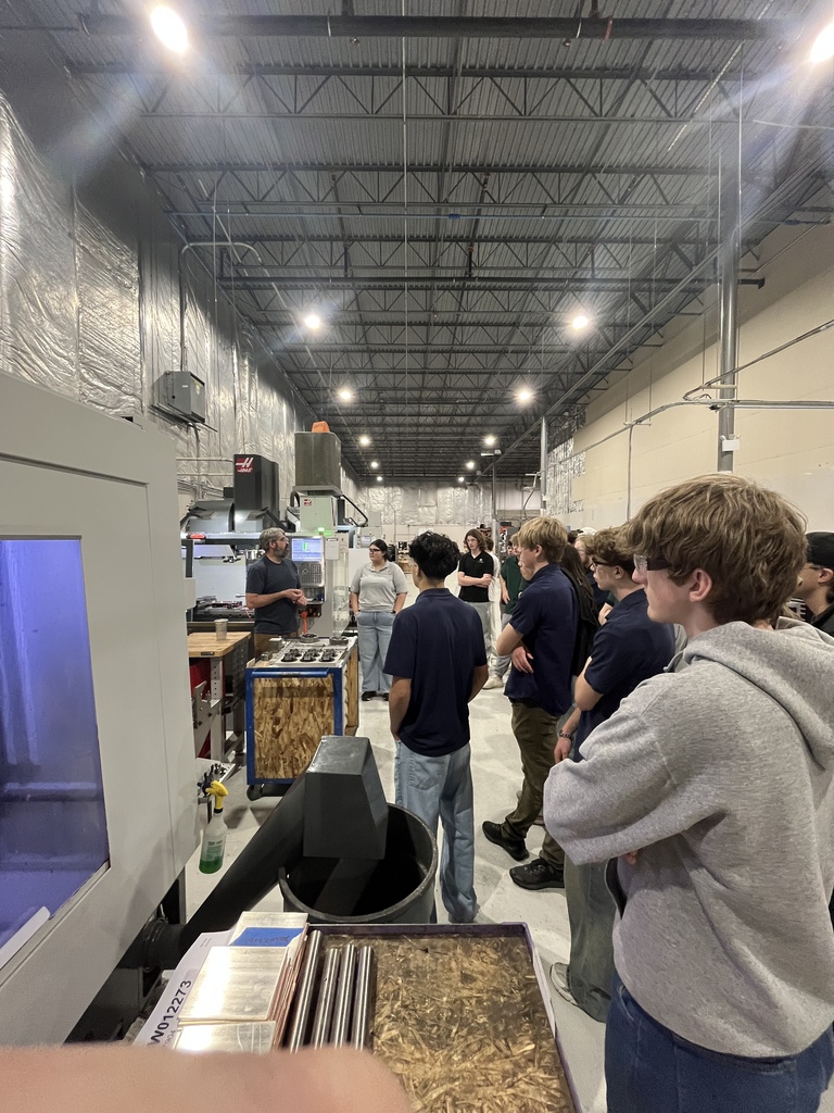 Students stand in a line watching an instructor speak near a large CNC machine inside a manufacturing shop. Worktables with metal parts and tools sit nearby, and the high-ceilinged industrial space contains multiple machines and bright overhead lights.
