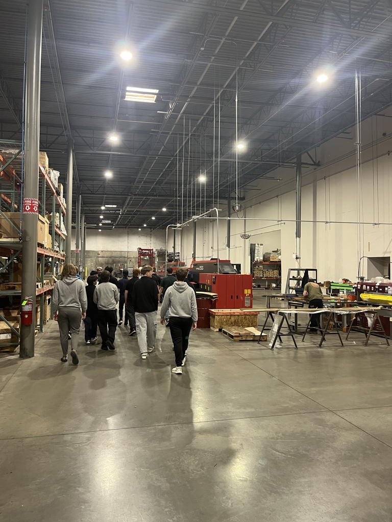 A group of students walks together through a spacious manufacturing warehouse with tall ceilings, bright overhead lights, and rows of shelving, worktables, and machines. Red industrial equipment and storage racks line the wide aisle as the group moves through the facility.