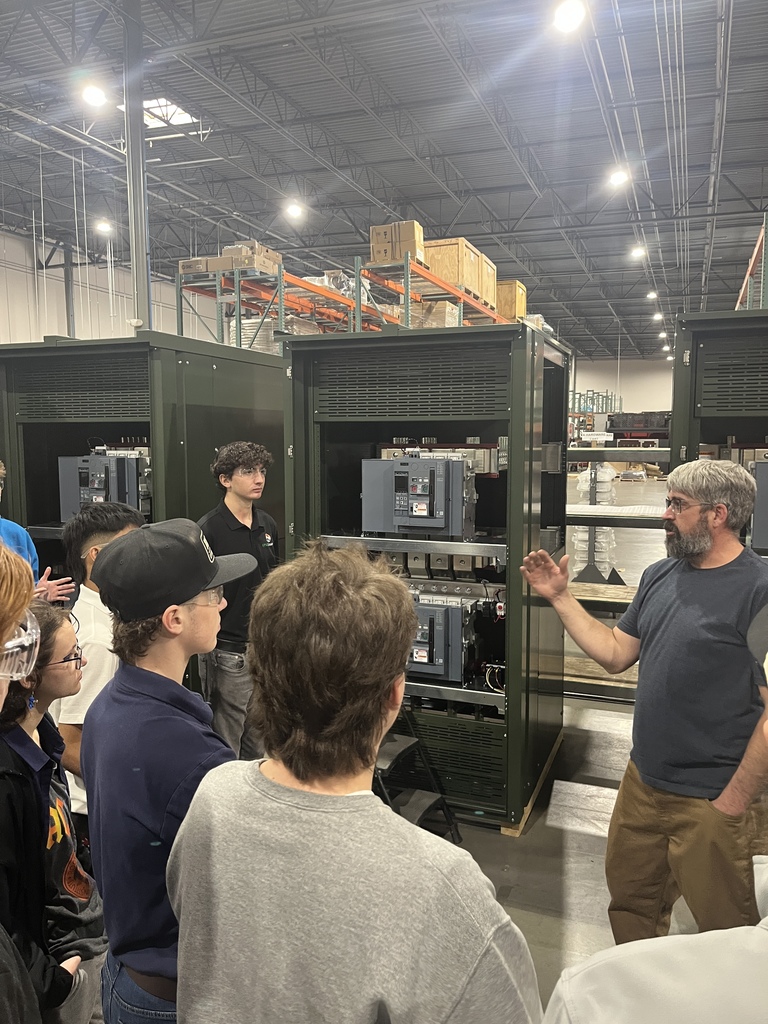 An instructor gestures while explaining large electrical control cabinets to a group of high school students standing nearby. The open cabinets reveal breakers and electrical components, and the group listens attentively in the industrial workspace.