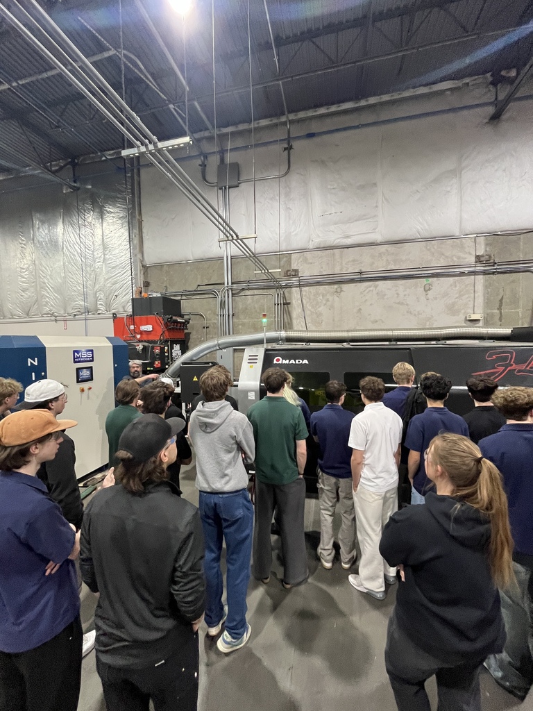 A group of students stands closely together observing a large industrial laser cutting machine labeled “Amada.” An instructor speaks from the side while the students watch the machine through its viewing window inside the busy manufacturing facility.