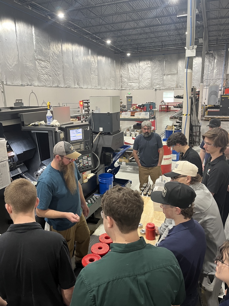 A group of high school students wearing safety glasses gathers around an instructor inside a large manufacturing facility. The instructor stands beside a CNC machine and explains a small metal part while students watch closely. Industrial equipment, tool holders, and worktables are visible around the workspace.