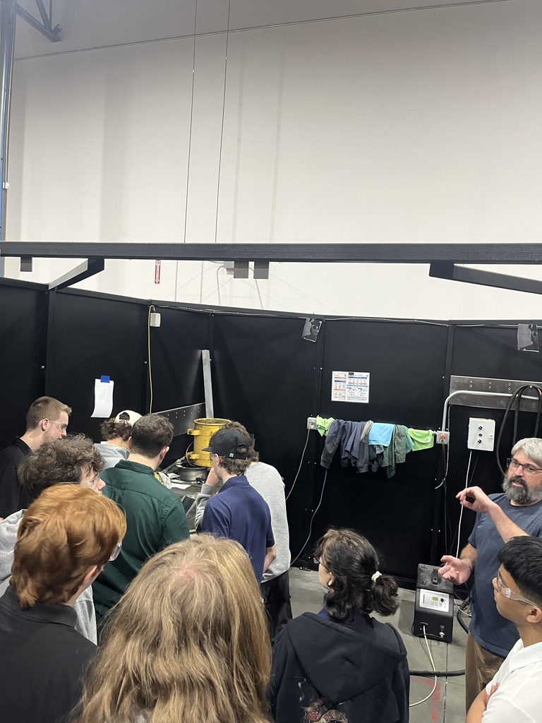 Students stand in a semicircle inside a workshop as an instructor gestures and explains a machining process near a workstation enclosed by black protective panels. The students, wearing casual clothing and safety glasses, observe the demonstration while equipment and cables line the walls.
