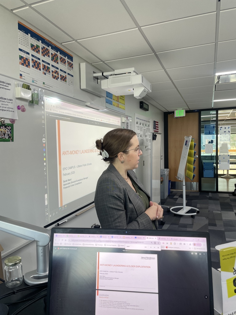 A guest speaker stands at the front of a classroom presenting a slide titled “Anti-Money Laundering & Elder Exploitation” for EPIC Campus–Littleton Public Schools. The presenter, wearing glasses and a gray blazer, speaks beside a projected presentation on a smartboard. Classroom items such as a periodic table chart, projector, and glass classroom doors are visible in the background.
