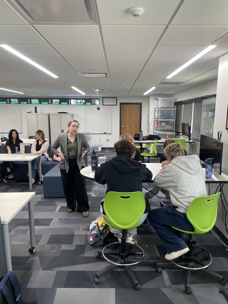 A guest speaker stands in the center of a modern classroom speaking with high school students seated at tables and green rolling chairs. A digital ticker-style display showing stock symbols such as “META” runs along the wall above whiteboards. Students listen and take part in the discussion in the EPIC Campus classroom.