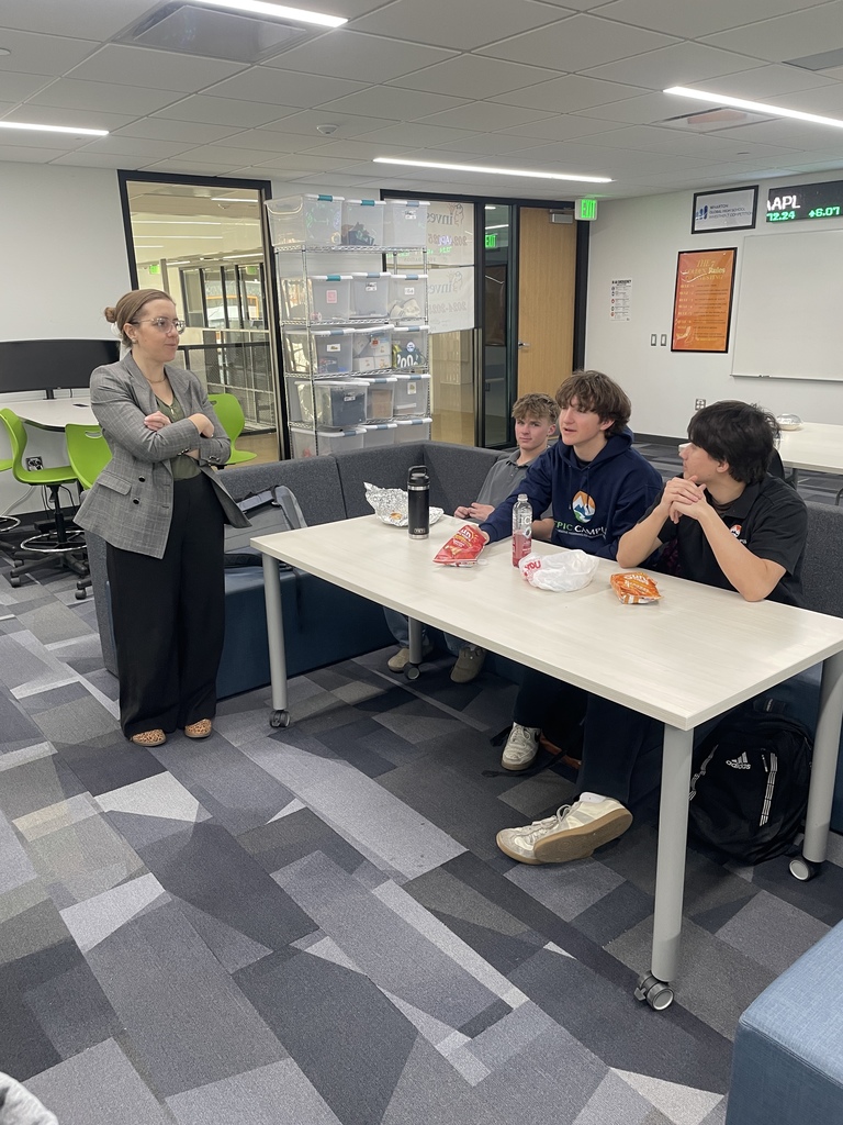 A guest speaker stands with arms crossed while talking with three high school students seated at a table in a collaborative classroom space. The students, wearing EPIC Campus apparel and casual clothing, listen while eating snacks. Storage bins, flexible seating, and glass-walled rooms are visible in the modern classroom environment.