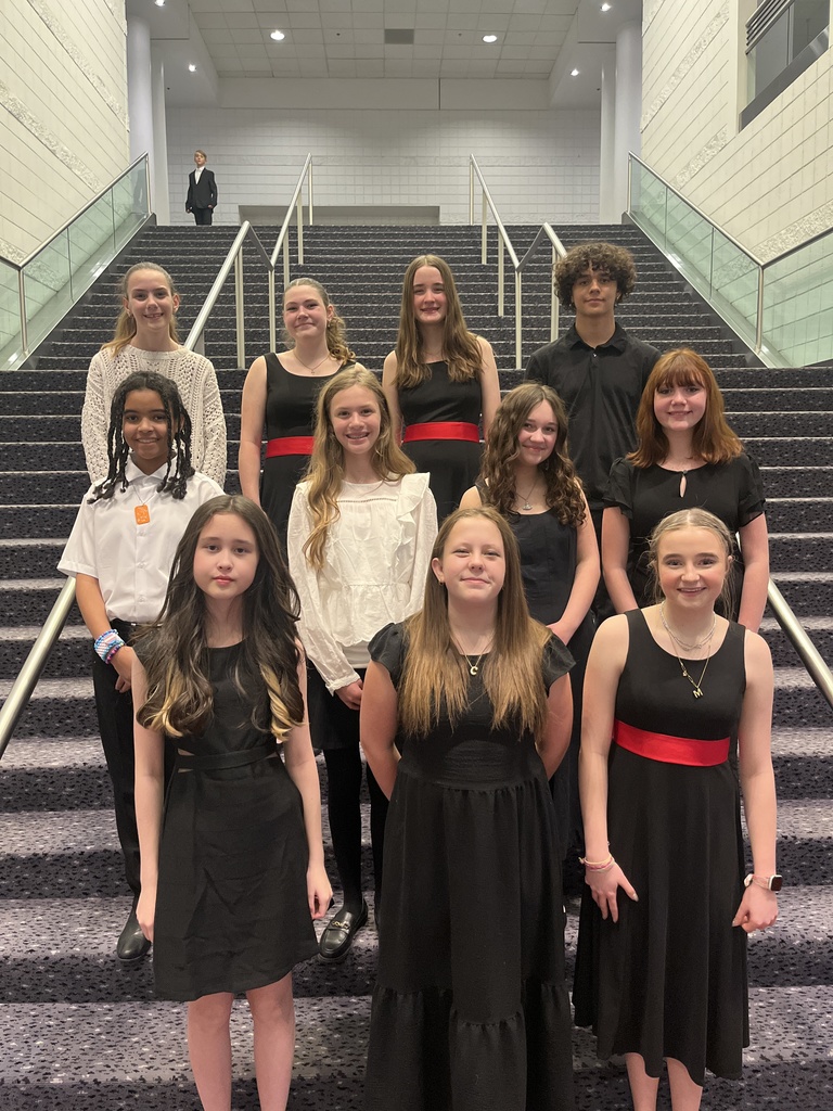A group portrait of ten middle school students standing on a wide, carpeted staircase in a theater lobby. The students are dressed in formal performance attire, primarily black dresses and shirts, with some wearing red sashes. They are smiling and posed in tiered rows.