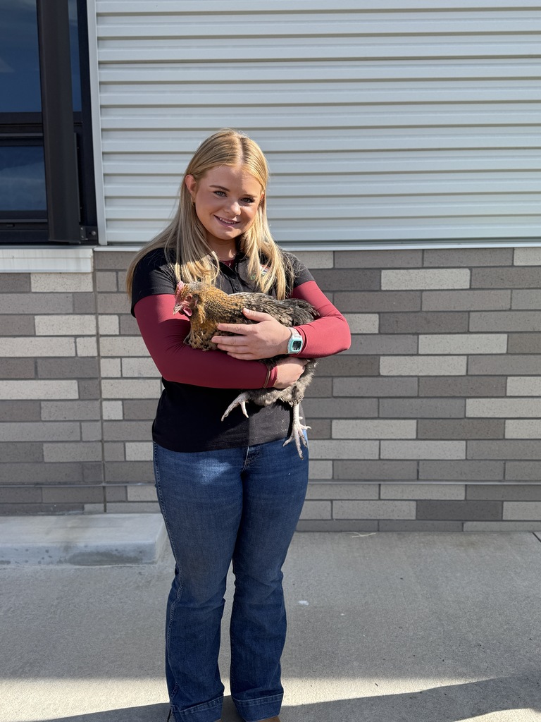 A smiling high school student stands outside a brick school building, holding a brown chicken in her arms.