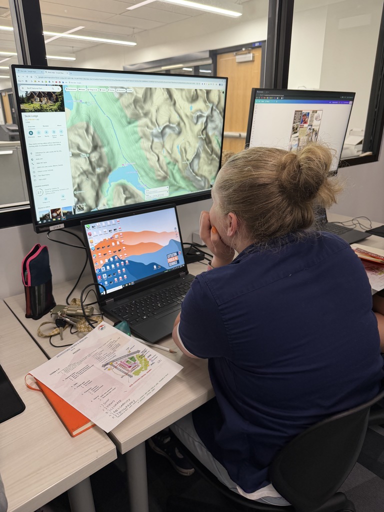 Student with hair in a bun sits at a workstation looking at a large monitor displaying a topographic map. A laptop, printed site plan with notes, keys, and a pencil case are arranged on the desk in a classroom setting.