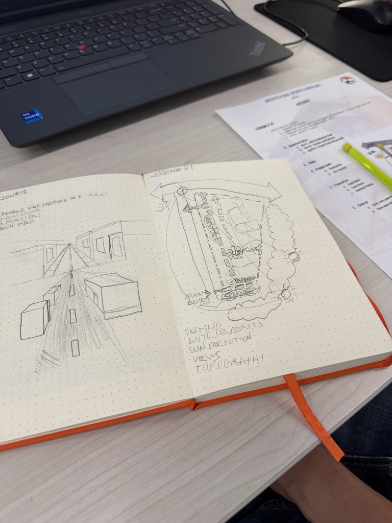 Open notebook on a desk shows detailed pencil sketches of building and site layouts, including labels for pathways, entries, and sun direction. A laptop and printed worksheet with a highlighted section sit nearby.