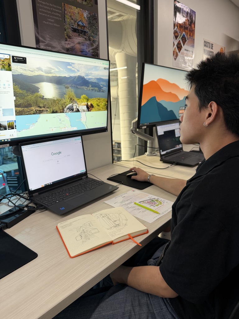 High school student sits at a desk using a multi-monitor computer setup, viewing a landscape and map on a large screen. A laptop displays Google, and a notebook with hand-drawn architectural sketches lies open beside printed planning materials.