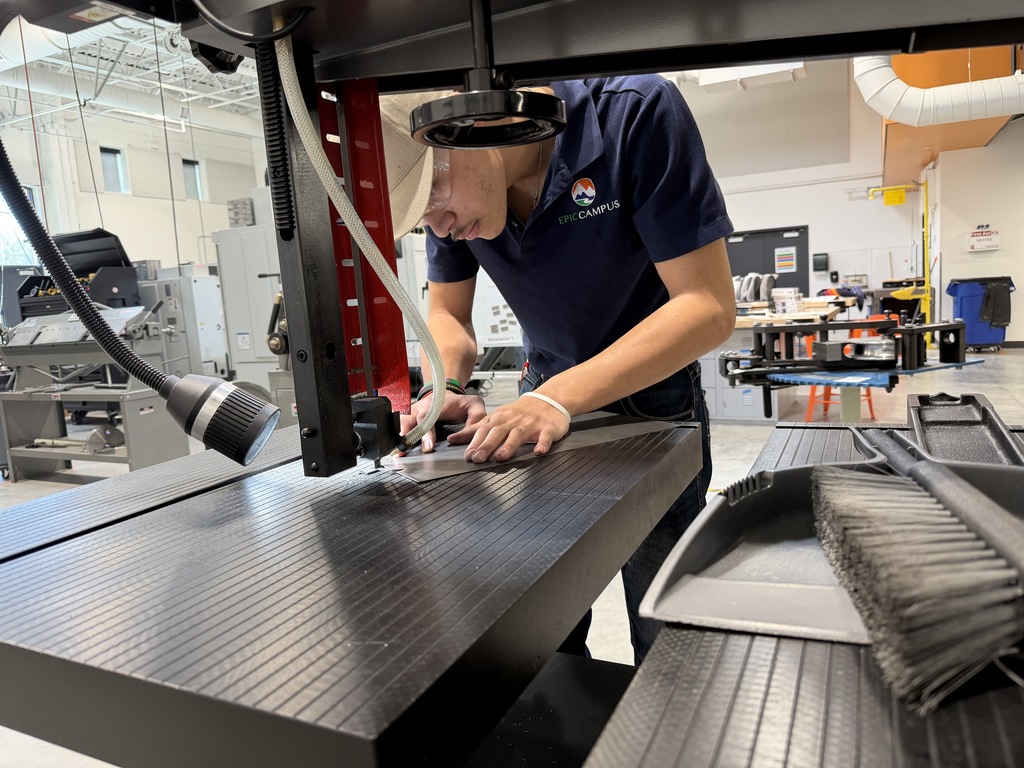 Student in an “EPIC Campus” shirt and cap uses a band saw to cut material, guiding it carefully with both hands. Workshop equipment and tools are visible in the background.