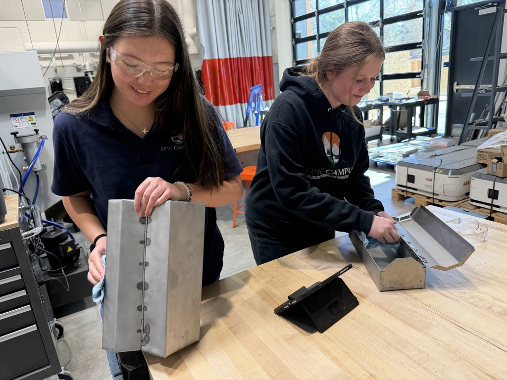 Two high school students in a workshop clean and handle metal fabrication pieces at a workbench. One student wears safety glasses and an “EPIC Campus” shirt; a tablet sits on the table nearby, and industrial equipment fills the background.