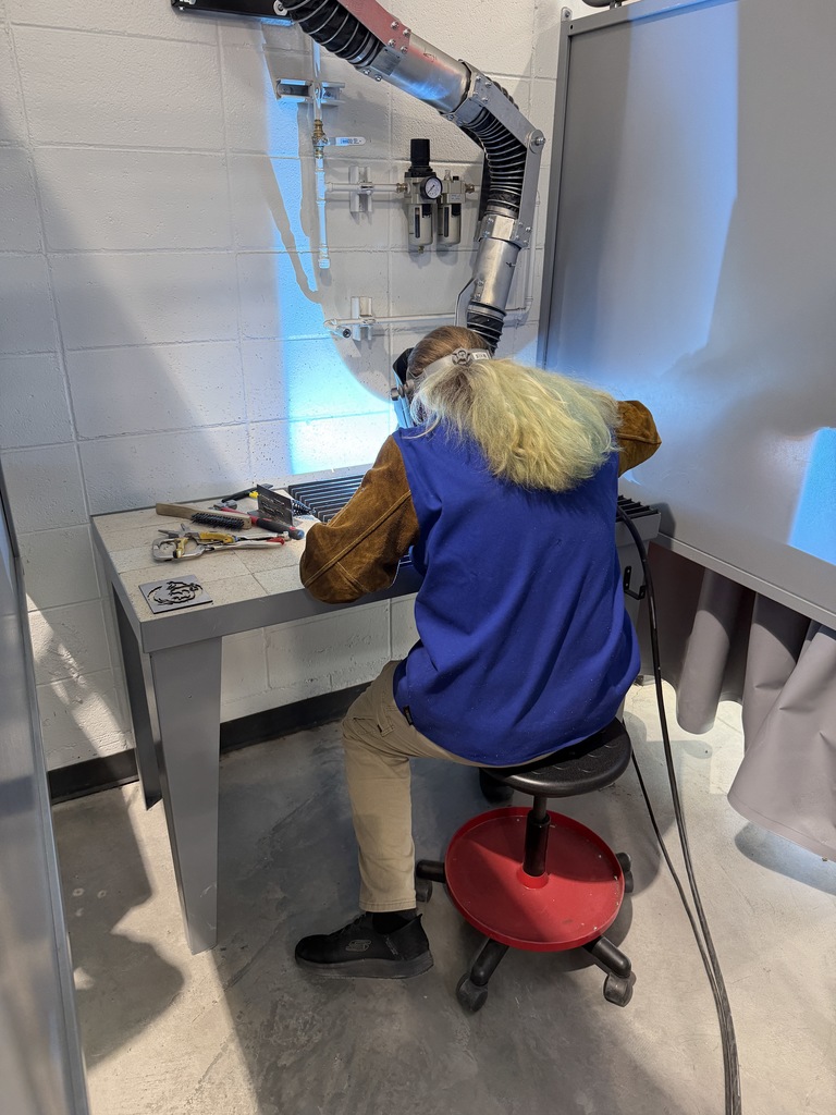 Student seated at a welding station wears protective gear and works at a metal table with tools nearby. A ventilation hood and piping system are mounted on the wall above the workspace.