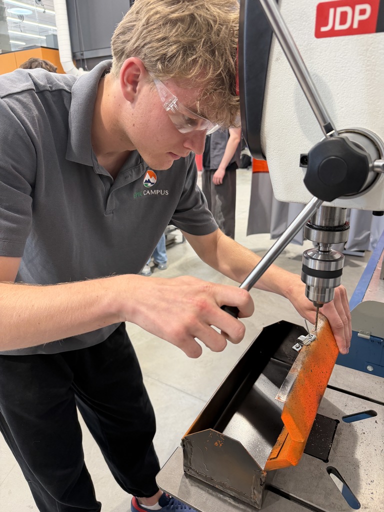 Student wearing safety glasses operates a drill press to fasten a hinge onto a metal project. The student focuses closely on the task in a manufacturing lab setting.