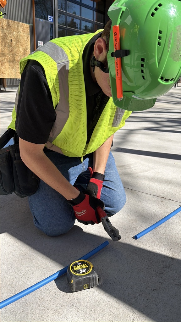 Student wearing a green hard hat, safety vest, and gloves kneels on concrete while cutting a blue conduit pipe with hand tools. A tape measure lies nearby.