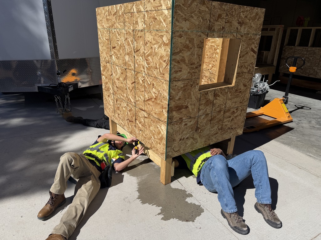 Two high school students wearing safety vests lie on the ground beneath a small wooden structure on wheels, using tools to fasten components underneath in an outdoor construction area.