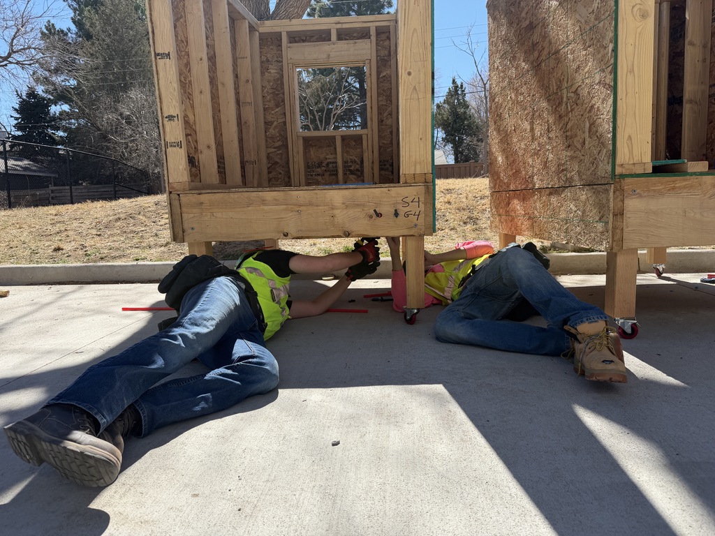 Two students in safety vests work underneath a small wooden structure, using tools to secure parts while lying on the ground. A pallet jack and workshop equipment are visible in the background.