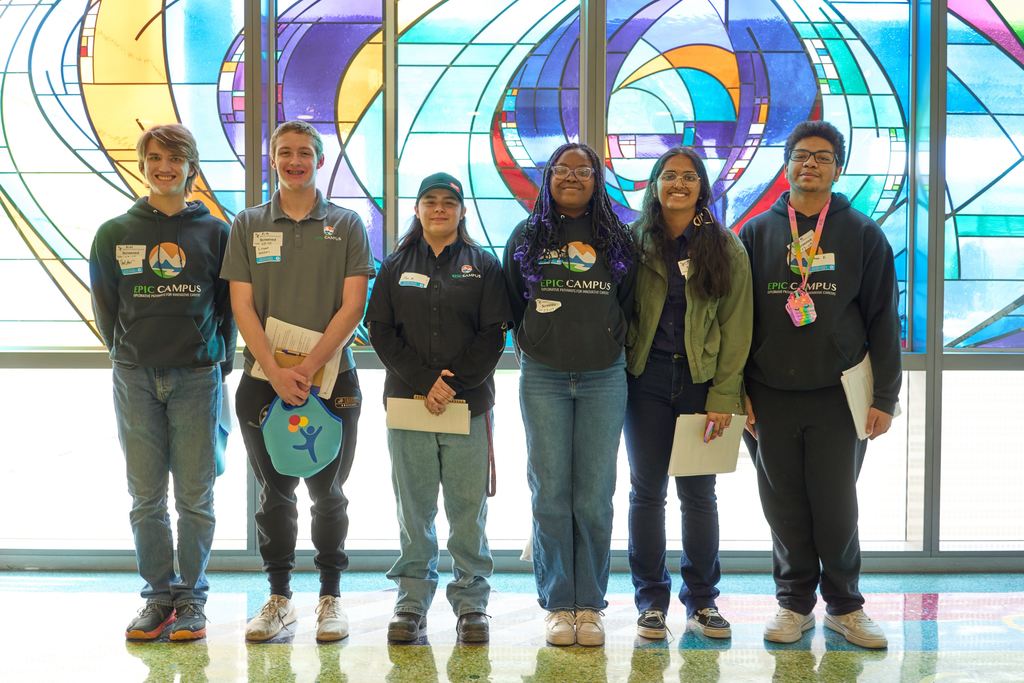 Six diverse high school students stand side by side indoors in front of a large, colorful stained glass window. Several wear “EPIC Campus” shirts or hoodies and name tags, and some hold notebooks or folders, smiling toward the camera.
