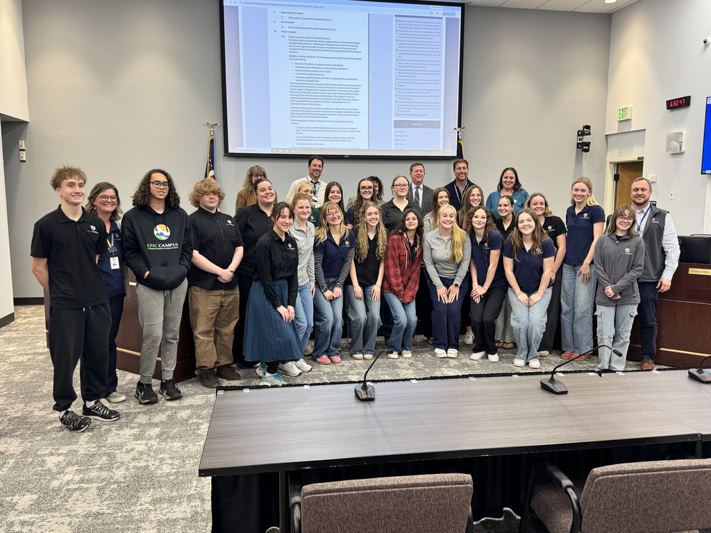 Large group of diverse high school students and several adults stand together at the front of a boardroom, smiling toward the camera. A presentation screen displaying a document is behind them, along with U.S. and state flags, while microphones and a conference table are visible in the foreground.