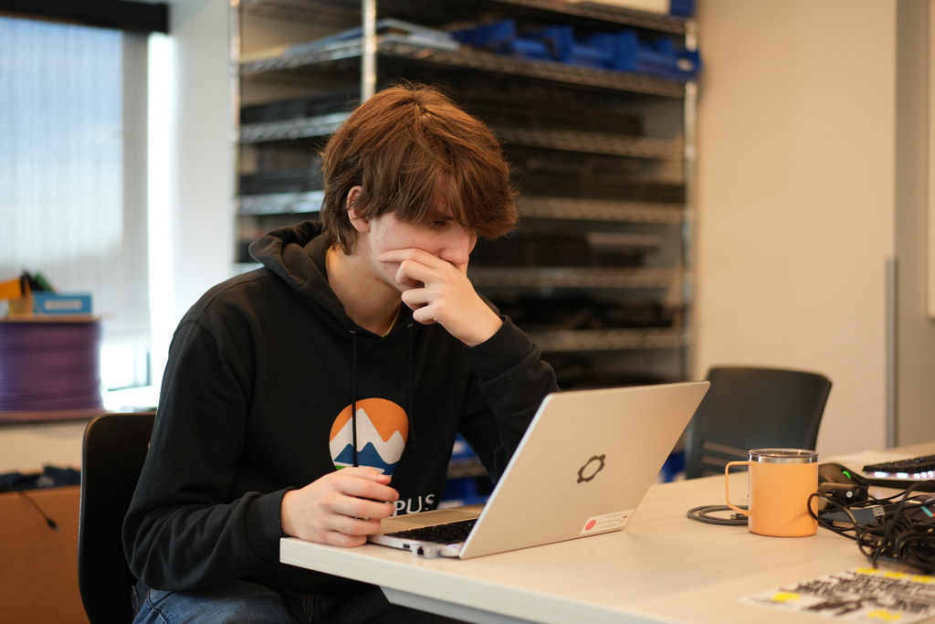Student in a black “EPIC Campus” hoodie works on a laptop, resting their hand near their face in concentration. A mug and coiled cables sit on the table beside them in a classroom workspace.
