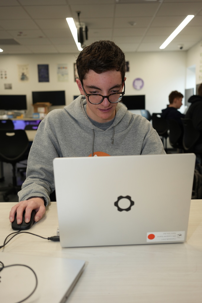 Student wearing glasses uses a laptop and mouse at a desk, focused on the screen in a classroom with other students working in the background.