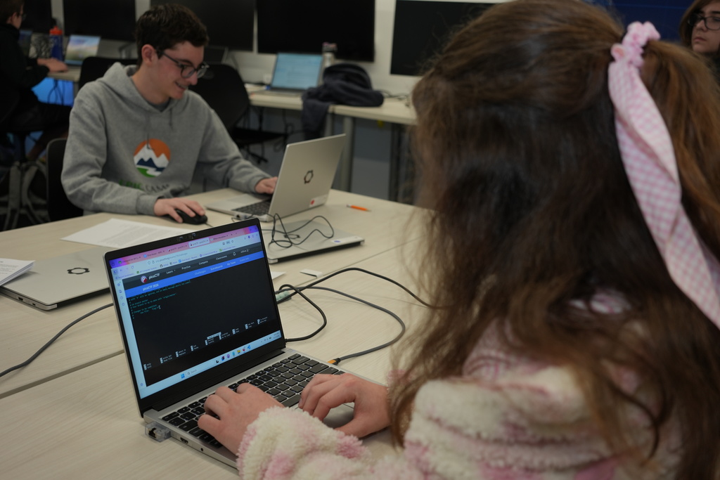 Student with long hair tied back types on a laptop displaying a coding interface, seated at a table with other students working on laptops in a computer lab.