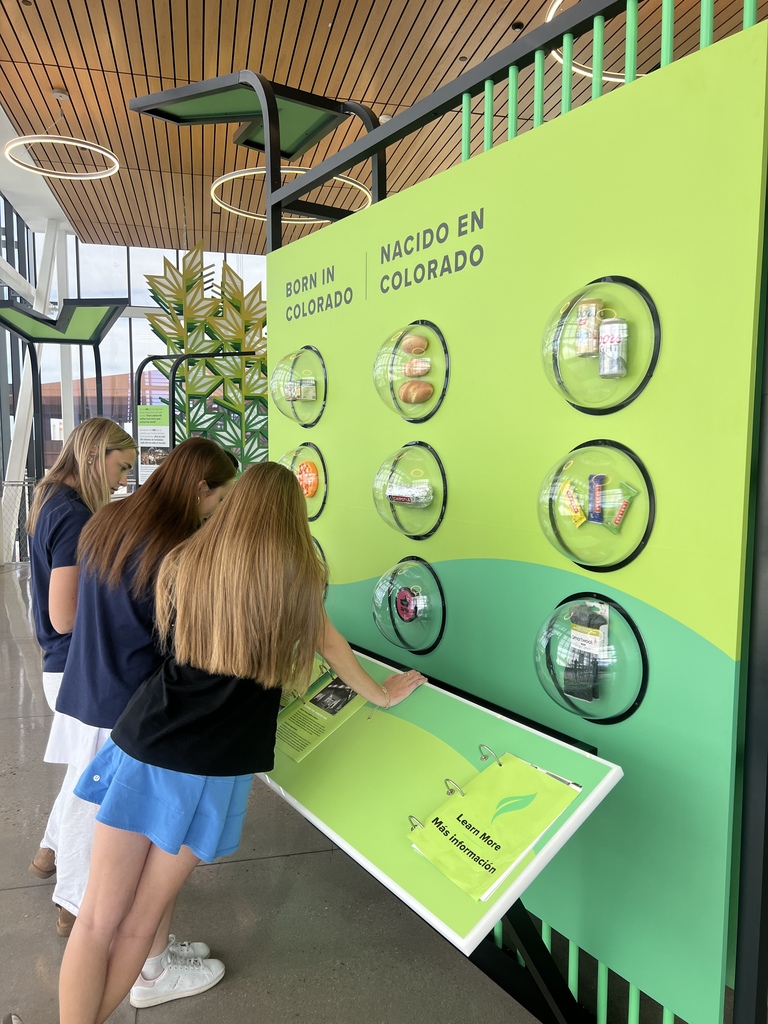 Three students read and interact with a bright green exhibit wall labeled “Born in Colorado / Nacido en Colorado,” featuring clear display domes with various Colorado-made products.