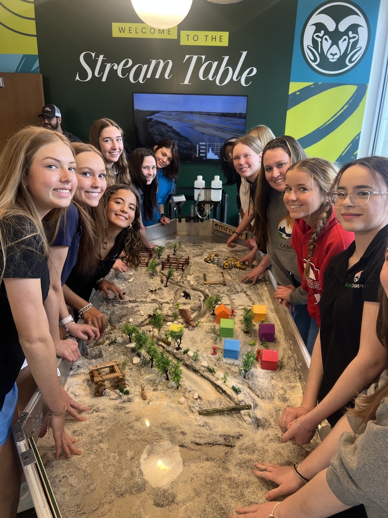 Group of students gathers around an interactive sand and water “stream table” model, smiling at the camera while exploring a miniature landscape with trees, buildings, and flowing water. A sign above reads “Welcome to the Stream Table.”