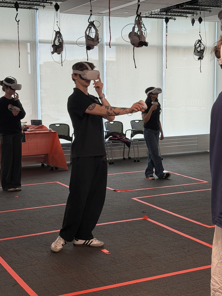 Students wearing virtual reality headsets stand in marked spaces on the floor, holding controllers and interacting with a VR experience in a classroom setting.